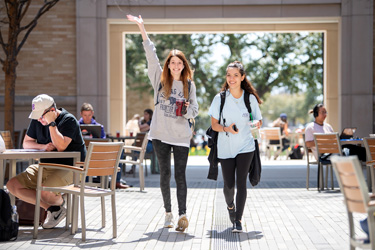 Plaza on a sunny day, with students at tables. Two students are walking and smiling at the camera.